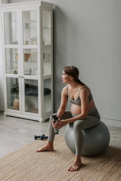 Full shot of young attractive expectant tired woman wipe the sweat from forehead with hand breathing and having rest sitting on fitness ball in living room at home. Healthy Pregnancy concept