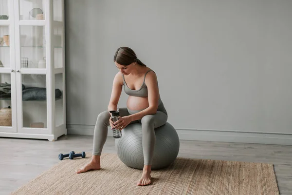 Full shot of young expectant woman with big belly in sportswear doing breathing and stretching exercises sitting on fitness ball in living room at home. Healthy Pregnancy concept 4K. High quality