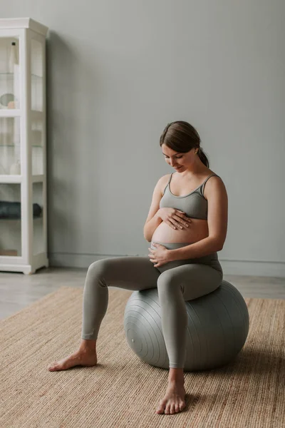 A pregnant woman in sportswear sits on a fitball and holds her hands on her stomach. Close-up view of arms and abdomen. Health care during pregnancy. Fitness for pregnant women.