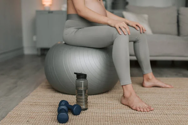 Full shot of young expectant woman with big belly in sportswear doing breathing and stretching exercises sitting on fitness ball in living room at home. Healthy Pregnancy concept