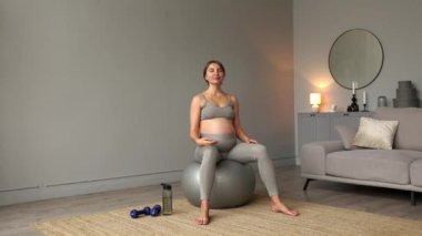 Spor kıyafetleri içinde koca göbekli, nefes alıp esneme egzersizleri yapan, evdeki oturma odasında fitness topuna oturan hamile bir kadın. Sağlıklı Hamilelik Konsepti 4K. Yüksek kalite 