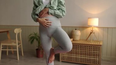 Rahat kıyafetler giyen hamile bir kadının evde oturma odasında tek ayak üzerinde yoga yaparken yakın plan fotoğrafı. İnsanlar ve hamilelik aktiviteleri konsepti. Sağlıklı ebeveyn kavramı. 4K. 