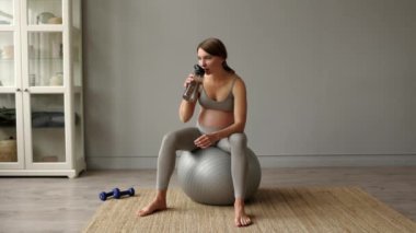 Genç, çekici, hamile, hamile bir kadın şişeden su içiyor ve evinde spor salonunda dinleniyor. Sağlıklı hamilelik konsepti 4K Yüksek kalite görüntü.