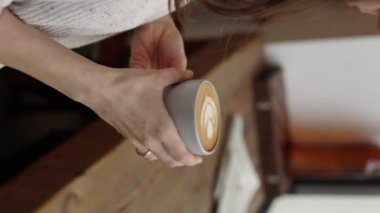 Çekici genç kadın ellerinin dikey pozu latte sanatıyla kahve tutuyor ve tavan arası Cafe 4k 'nin penceresinin önündeki ahşap masanın yanında keyfini çıkarıyor. Yüksek kalite 4k görüntü