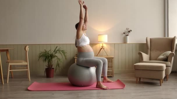 午前中は自宅のリビングルームでフィットネスボールに座って練習を行う魅力的な笑顔妊娠中の女性の側面図 幸せな健康的な妊娠の概念4K 高品質4K映像 — ストック動画