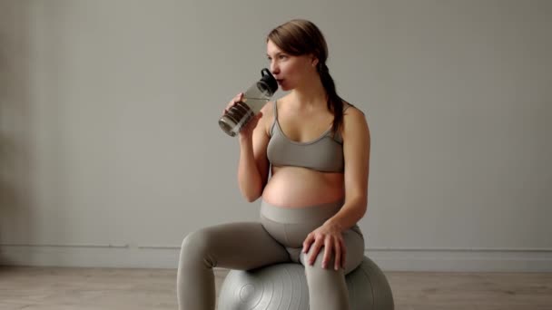トレーニングや飲料水の後 フィットネスボールに座って穏やかな妊婦 期待の母親のフロントビューのリビングルームで自宅で休んでいる 妊娠コンセプト4K 高品質4K映像 — ストック動画