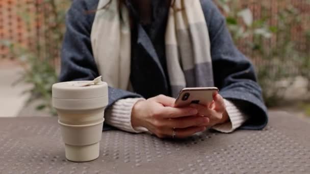 在喝咖啡休息期间 坐在室外用手机发短信或冲浪的无法探测的女性近照 带着生态可重复使用的杯子 在市中心或公园边穿着带围巾4K的外套 — 图库视频影像