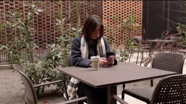 エコ再利用可能なコーヒーブレイク中に屋外に座っている携帯電話で幸せな女性のテキストメッセージやサーフィンは スカーフ4Kとコートで市内中心部や公園側にカップを維持します 高品質4K映像 — ストック動画