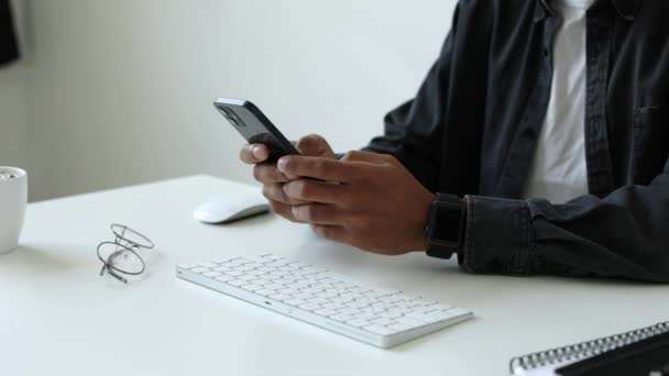 認識されていないアフリカ系アメリカ人の中程度のショットスマートフォンデバイスを保持している人のテキストメッセージミリ秒は ソーシャルメディアでチャットアプリプレイモバイルゲームを使用して自宅のオフィスでキーボードで白い机に座っている — ストック動画