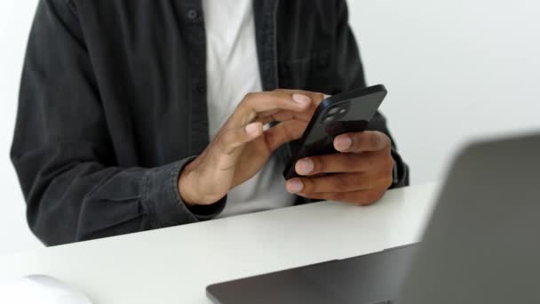 在现代公寓或办公室里用智能手机坐在白桌旁的深色皮肤男人的近照 发短信 滚动点击技术生活方式 通过电话远程工作4K — 图库视频影像
