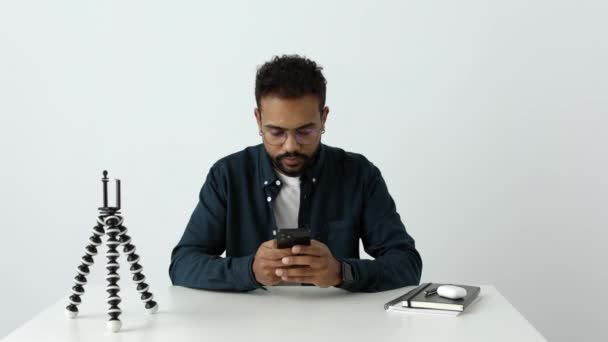 戴眼镜的快乐的黑人绅士坐在家里的写字台旁 在智能手机上浏览社交媒体 美国电话用户通过手机博客4K输入短信阅读电子邮件 — 图库视频影像