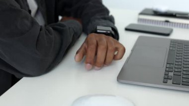 Siyah tenli bir adamın eli, tedirgin bir şekilde beyaz bir masaya dokunurken, dizüstü bilgisayarın önünde, teknolojik aletlerle otururken yakın plan görüntüsü.