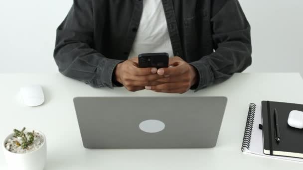 白い風通しの良いオフィスでノートパソコンの前に座っているスマートフォンにアフリカ系アメリカ人男性を入力し テキストメッセージの高い角度ビュー 屋内黒大人の学生オンライン研究 暗号愛好家検索株式 — ストック動画