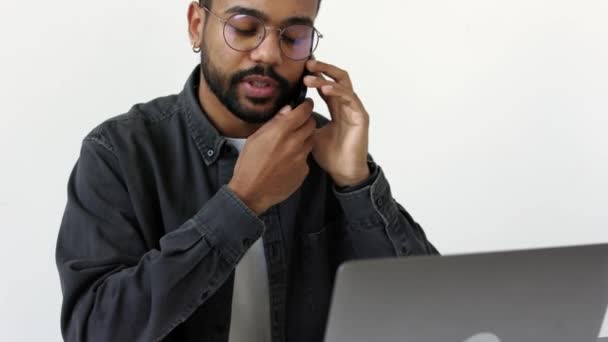 深刻な黒肌の男で眼鏡で灰色のシャツの話で携帯電話を保持スマートフォンでシェルと頭によって光オフィススタジオでノートパソコンやテキストメッセージやブログの前に座って4K — ストック動画