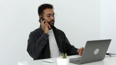 Yandan gülümseyen, şık gri gömlekli, kıvırcık saçlı, gözlüklü, laptopta çalışan ve telefonda konuşan havalı, rahat modern ofis stüdyosu 4k 'te beyaz masada oturan mutlu Afrikalı bir erkek..