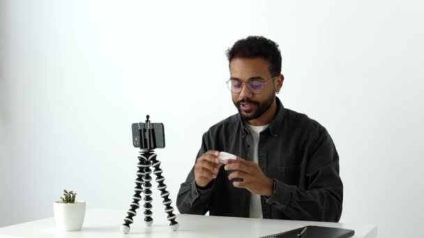 幸せな若いスマートブラックデジタルブロガー男性と眼鏡撮影新しいVlogビデオとともに三脚カメラ電話で自宅オフィス プレゼンテーションやイヤフォンの新しいタイプのボクシング高品質4K映像 — ストック動画