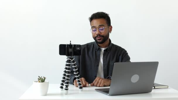 一位英俊的非洲裔美国男子坐在白色桌子边 手持笔记本电脑 头戴绿色的三脚架 在为他的网络追随者拍摄的最新博客中对着摄像机讲话 — 图库视频影像