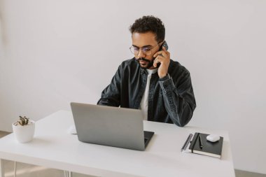 Gözlüklü, gri tişörtlü, kıvırcık saçlı, ciddi siyahi erkek. İş arkadaşlarıyla cep telefonuyla konuşuyor. Beyaz, temiz bir masada oturuyor. 