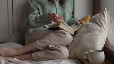 Yakın plan kadın ya da kız kitap okuyor balkon penceresinin yanında yastıkla oturuyor ve sıcak çay ya da kahve içiyor. Yüksek kalite 4k görüntü