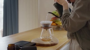 V60 'la kahve içerken demleme işlemi. Barista kızı ev mutfağındaki kağıt filtreye sıcak suyla toprak tohumu döküyor. Yüksek kalite 4k görüntü