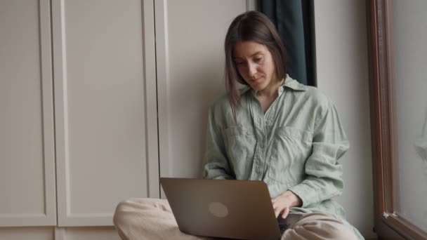 年轻漂亮的女人坐在笔记本电脑上打字 双腿交叉地坐在靠近卧室窗户的地板上 高质量的4K镜头 — 图库视频影像