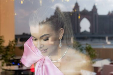 Çok güzel bir kız çiçeklerin arasında pencerede gülümsüyor. Kadınların mutluluğu. Çekici bir kişilik. Camdaki yansımalar. Olumlu ruh hali buketi. Tatil portresi. Pencere camından bakıyorum.