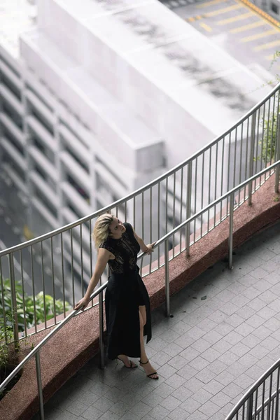 A beautiful Caucasian blonde girl in a luxurious black dress is standing against the backdrop of a park in a modern city. Greenery and concrete in the city of the future. High quality photo