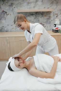 Kozmetik uzmanı, spa merkezinde bir sedyede yatarken kadına yüz masajı yapıyor. Rahat genç bayan kanepede dinleniyor.