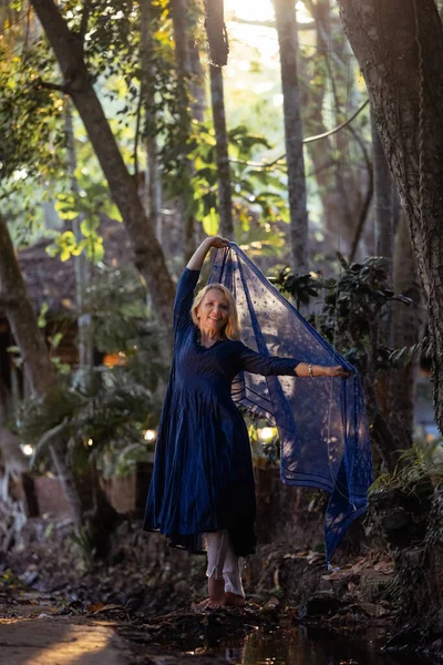 A beautiful fifty-year-old woman with light hair, dressed in a blue dress, is looking into the frame. The scene around her consists of nature during sunset.