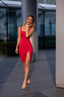 Kırmızı elbiseli, külotlu çoraplı ve topuklu ayakkabılı güzel sarışın kız şehir meydanında poz veriyor. Yüksek kalite fotoğraf