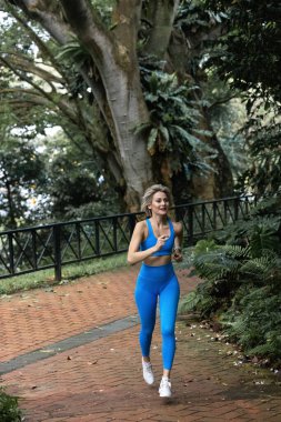 Güzel, zayıf bir sarışın büyük bir şehirde yeşil bir parkta mavi taytlı koşuyor. O gülümsüyor. Kardiyo çalışmaları önemlidir. - Evet. Yüksek kalite fotoğraf