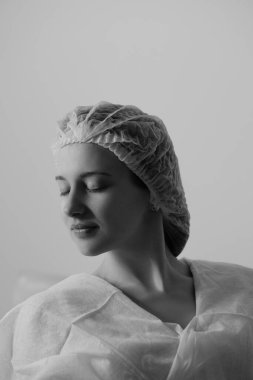 Tek kullanımlık tıbbi bir kep ve hasta önlüğü takan genç bir kadının siyah beyaz fotoğrafı. Klinik ortamda sakin ve ciddi bir atmosfer. Yüksek kalite fotoğraf