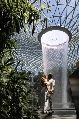  Singapur 'daki Jewel Changi Uluslararası Havalimanı' nda yapılan düğün öncesi fotoğraf çekimi sırasında gelinlik giymiş beyaz takım elbiseli Asyalı bir gelin ve çeşmelerin yanında duran Asyalı bir damat. Yüksek kalite fotoğraf