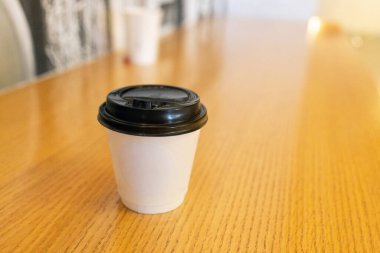 Sarı ahşap bir masanın üzerinde siyah plastik kapaklı beyaz kahve fincanı. Minimalist tarzda ve sıcak tonlarda. Bir kafede ya da açık havada sıcak bir kahve molası anı. Yüksek kalite fotoğraf