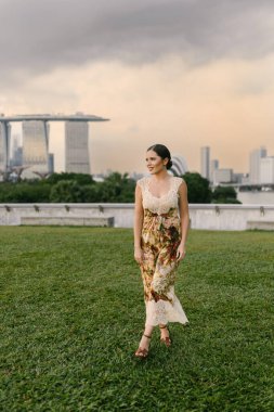 Marina Bay Sands Singapur siluetine karşı poz veren, çiçekli kum rengi bir elbise giymiş güzel Doğu Avrupalı ya da Kafkas bir kadın modern mimari ve şehir atmosferiyle. Yüksek kalite fotoğraf