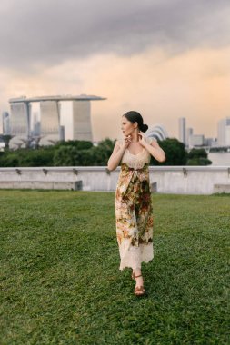 Marina Bay Sands Singapur siluetine karşı poz veren, çiçekli kum rengi bir elbise giymiş güzel Doğu Avrupalı ya da Kafkas bir kadın modern mimari ve şehir atmosferiyle. Yüksek kalite fotoğraf