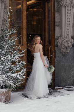 Taze çiçek buketiyle güzel bir gelin karlı arazide, kır ağacının yanında poz veriyor. Soğuk hava, şık elbise, şenlikli atmosfer ve romantik kış havası. Yüksek kalite fotoğraf