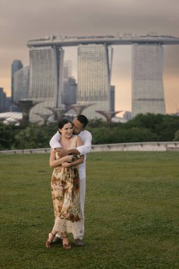 Singapur 'da Marina Bay Sands' in önünde kucaklaşan uluslararası bir çiftin yakın çekimi. Asyalı erkek ve Doğu Avrupalı kadın açık havada romantik bir an paylaşıyorlar, şık ve duygusal bir ortam. Yüksek kalite fotoğraf