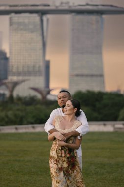 Singapur 'da Marina Bay Sands' in önünde kucaklaşan uluslararası bir çiftin yakın çekimi. Asyalı erkek ve Doğu Avrupalı kadın açık havada romantik bir an paylaşıyorlar, şık ve duygusal bir ortam. Yüksek kalite fotoğraf