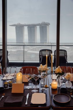Marina Bay Sands ve Singapur ufuk çizgisini birinci elden gören şık bir yemek düzeni. Rafine sofra takımı ile lüks restoran ortamı, sofistike bir şehir deneyimi sunuyor..