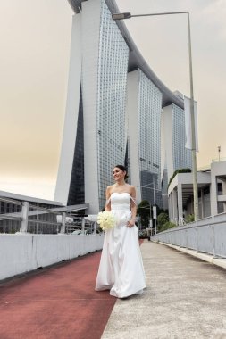 Şık beyaz gelinlikli neşeli gelin canlı bir şehirde çağdaş minimalist binaların arka planına poz veriyor. Şehir zarafeti ve düğün güzelliği açık havada yakalanmış..