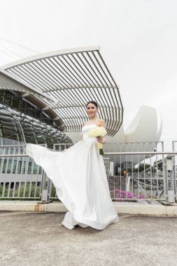 Şık beyaz gelinlikli neşeli gelin canlı bir şehirde çağdaş minimalist binaların arka planına poz veriyor. Şehir zarafeti ve düğün güzelliği açık havada yakalanmış..