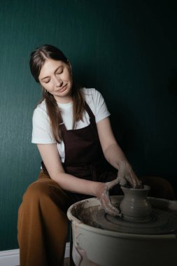 Koyu saçlı neşeli kadın seramik stüdyosunda çömlekçilik hobisi yapıyor. Elleri kille kaplı, çanak çömlek aletleri ve ekipman kullanarak el yapımı kil eşyalar üretiyor..