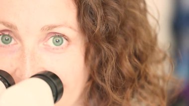 Klinikteki tıbbi mikroskoba bakarken gülümseyen kızıl saçlı kadın doktor. Yüksek kalite 4k görüntü