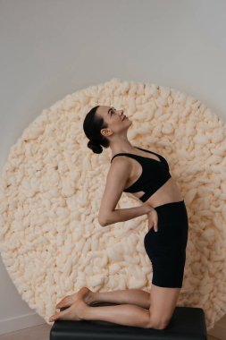 Kafkasyalı ince esmer kadın, duvarın arka planında büyük yün örülmüş kumaşla, rahat stüdyo içinde yoga asanas yapıyor.