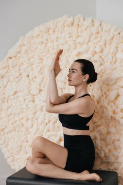 Kafkasyalı ince esmer kadın, duvarın arka planında büyük yün örülmüş kumaşla, rahat stüdyo içinde yoga asanas yapıyor.