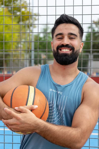 Basketbol topu tutan ve şehir basketbol sahasındaki kameraya bakan beyaz bir genç.