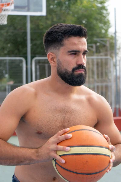 Basketbol topu tutan ve şehir basketbol sahasındaki kameraya bakan beyaz bir genç.