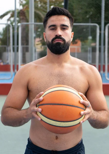 Basketbol topu tutan ve şehir basketbol sahasındaki kameraya bakan beyaz bir genç.