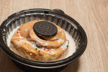 Beyaz kremayla kaplı tarçınlı tarçınlı börek ve üzeri çikolatalı sandviç kurabiyesi ve ahşap bir masada duran siyah bir kasenin içine serpiştirilmiş krema.